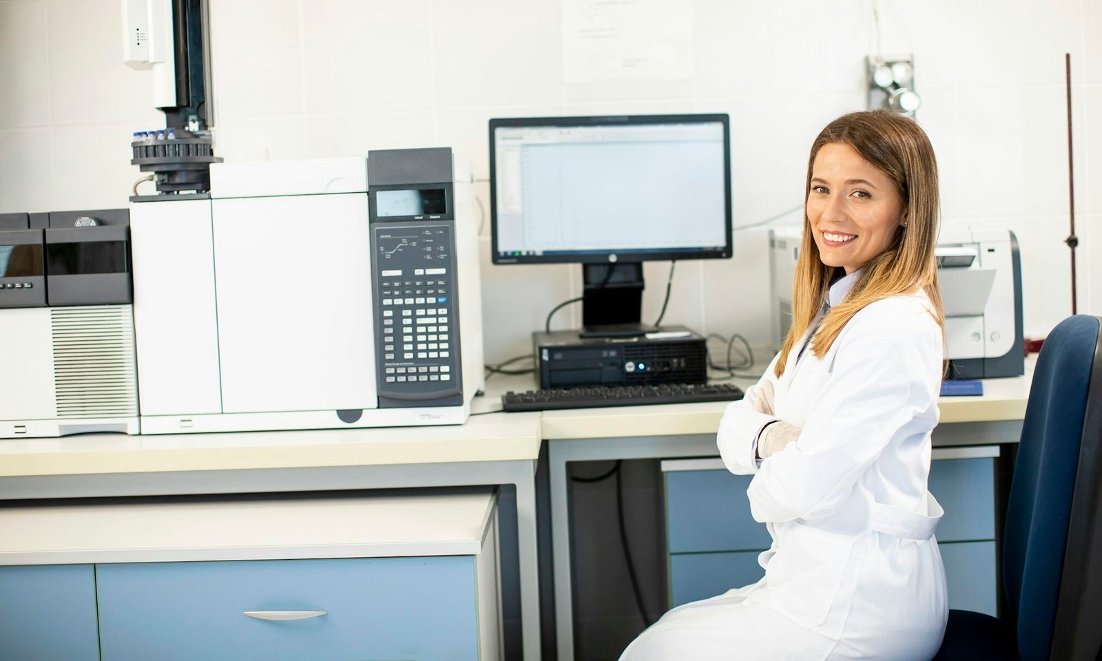 Female scientist in white lab coat sitting by the gas chromatography equipment in the biomedical lab