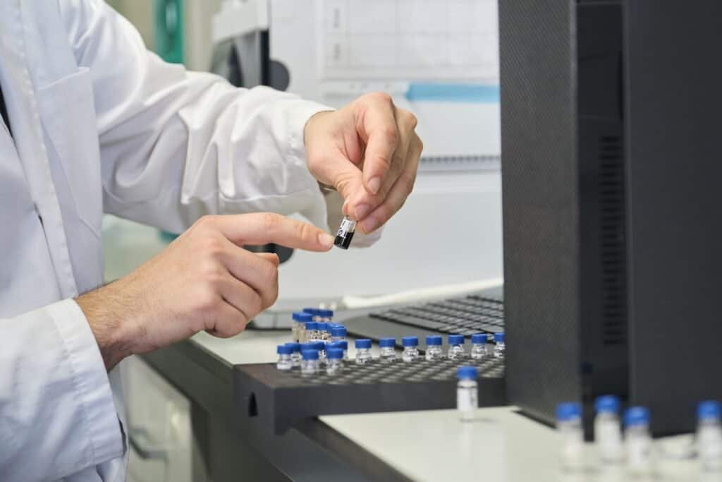 Scientist in lab coat putting vial with sample into autosampler of HPLC system.