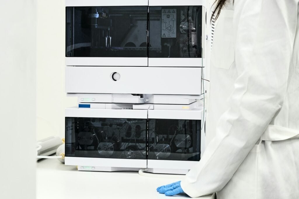 Scientist in rubber gloves and white lab coat standing near high performance liquid chromatography