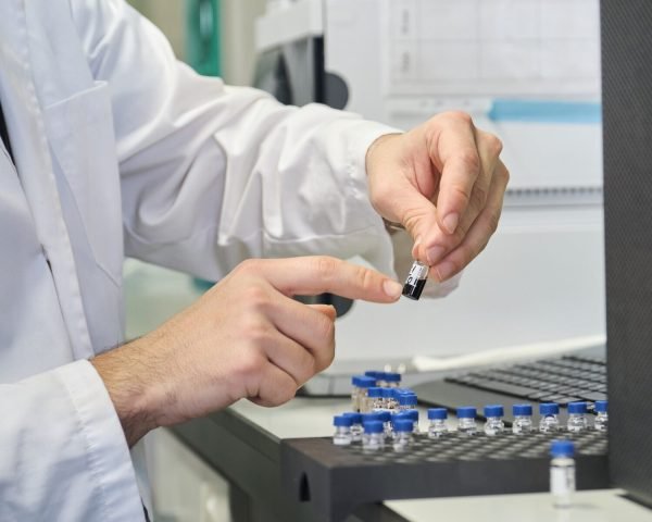 Scientist in lab coat putting vial with sample into autosampler of HPLC system.